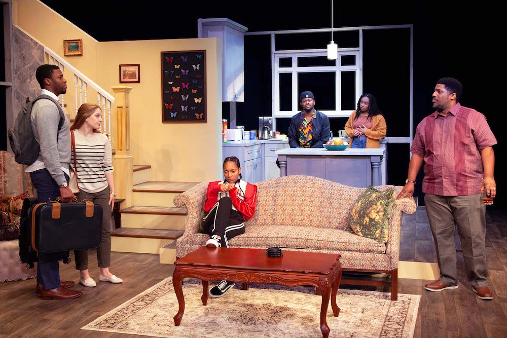 A cast of actors performs on a set that looks like a living room. 