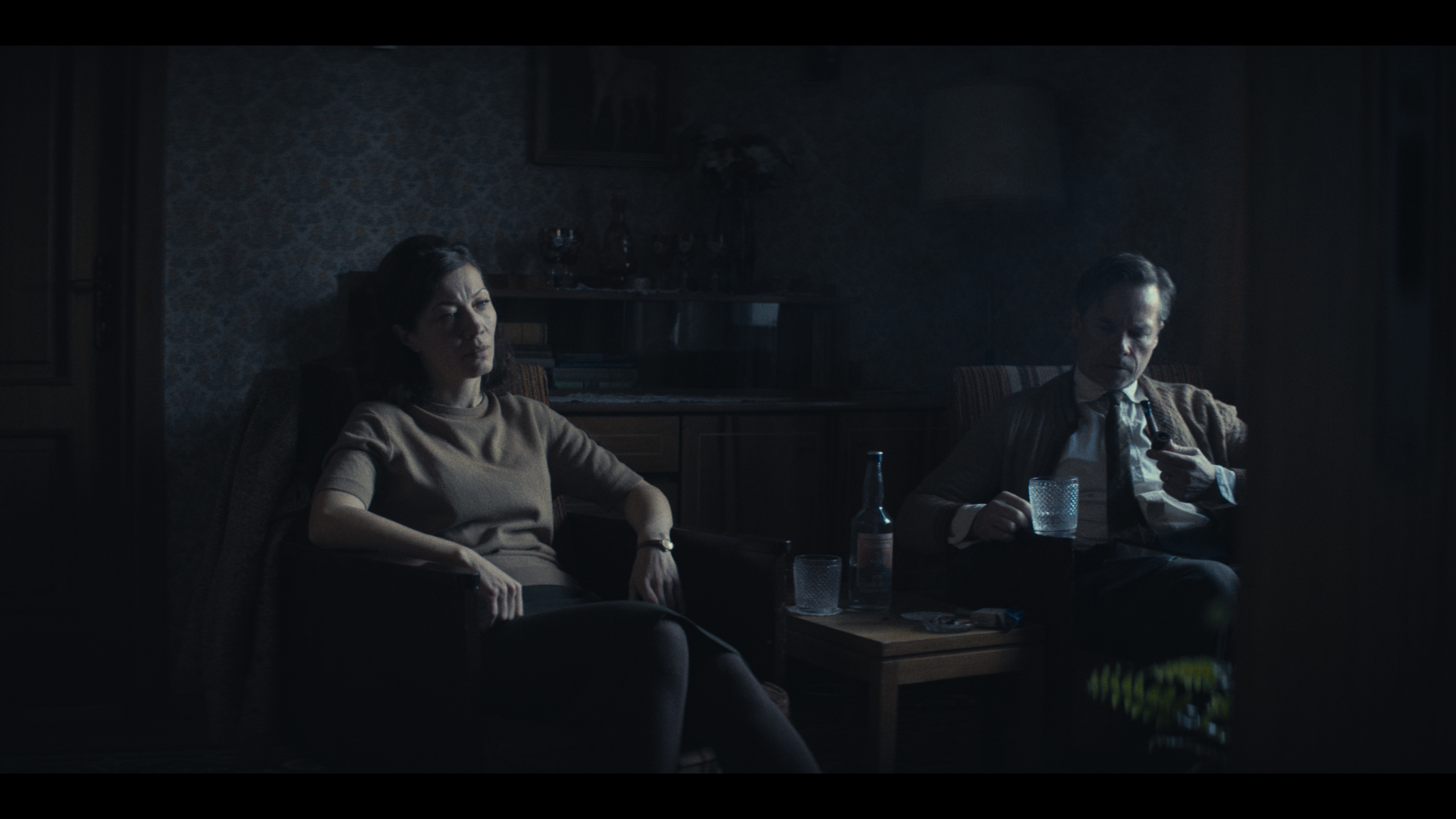 Two people sit in chairs in a darkly lit room. 