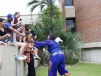 An actor in a purple costume runs towards a laughing audience in the FAB courtyard.