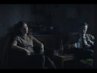 Two people sit in chairs in a darkly lit room.