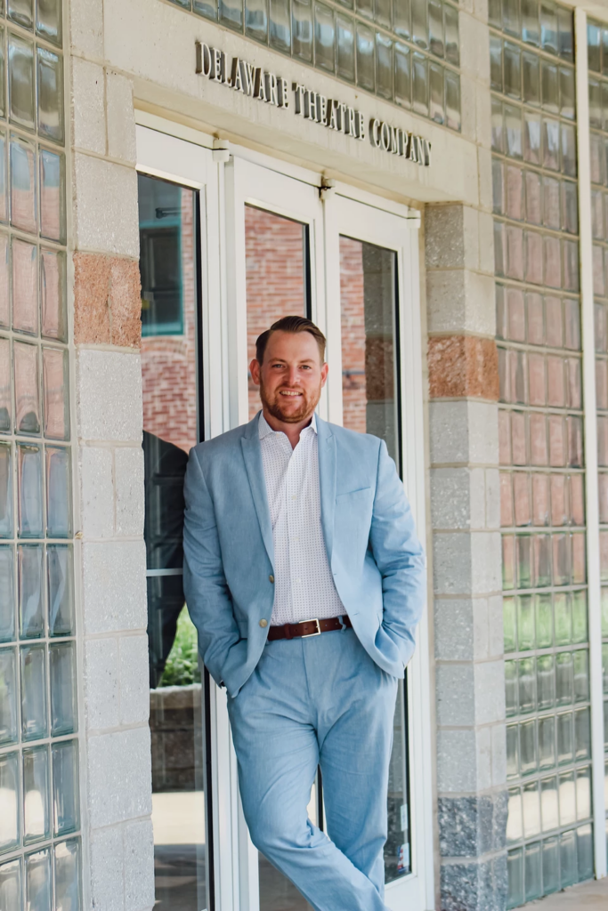 Matt Silva is wearing a light blue suit and leaning against a door under a sign that reads Delaware Theatre Company