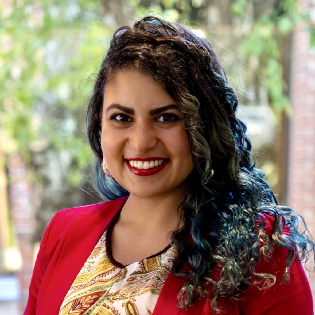 A woman with dark wavy hair smiles at the camera for a professional headshot. She is wearinf a paisley top and a red blazer. 